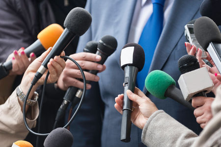 Multiple reporters holding microphones around a person in a suit and blue tie, conducting an interview.