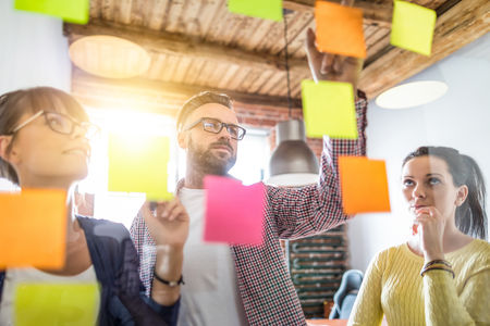 Three people, two women and a man, stand in front of a glass wall covered with colorful sticky notes, engaged in discussion and planning.