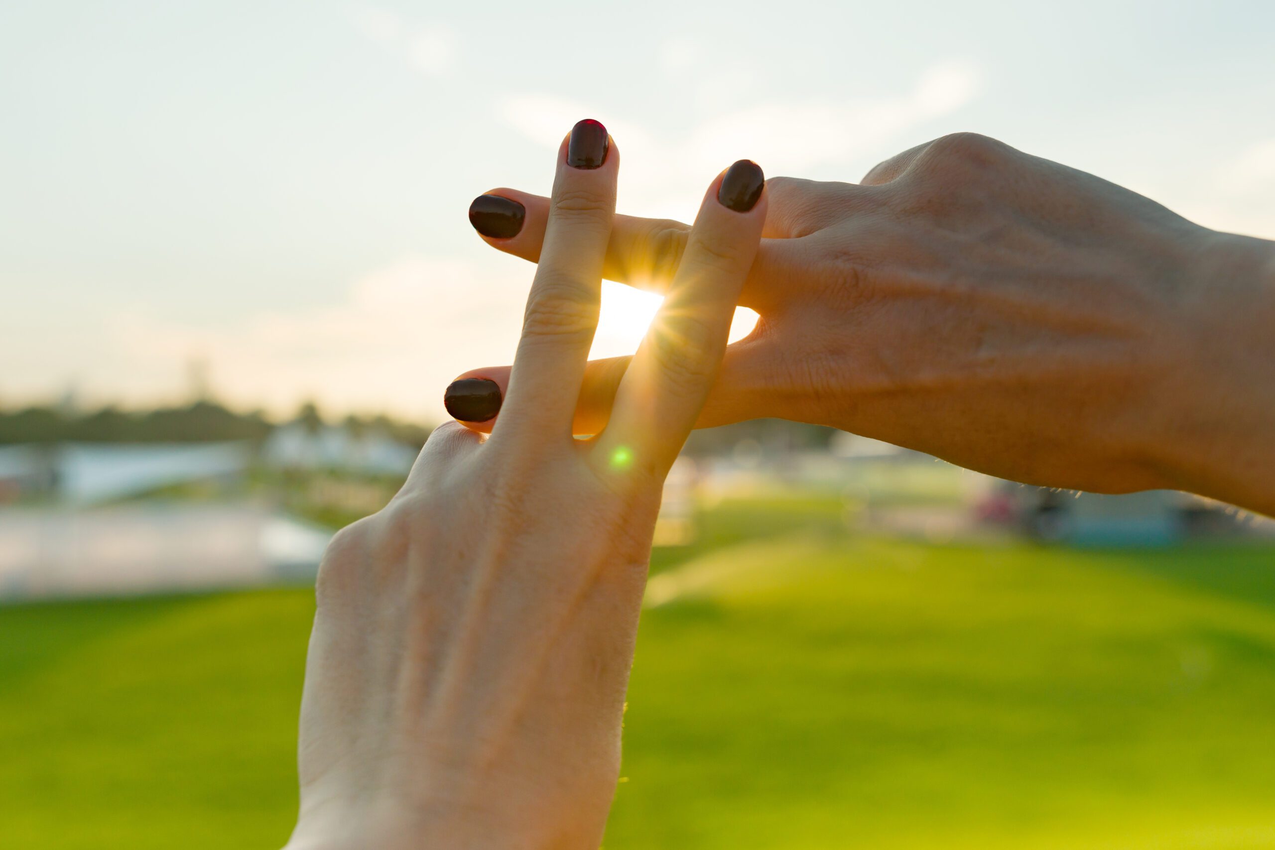 Two hands with painted nails form a hashtag symbol against a backdrop of green grass and a bright sky.