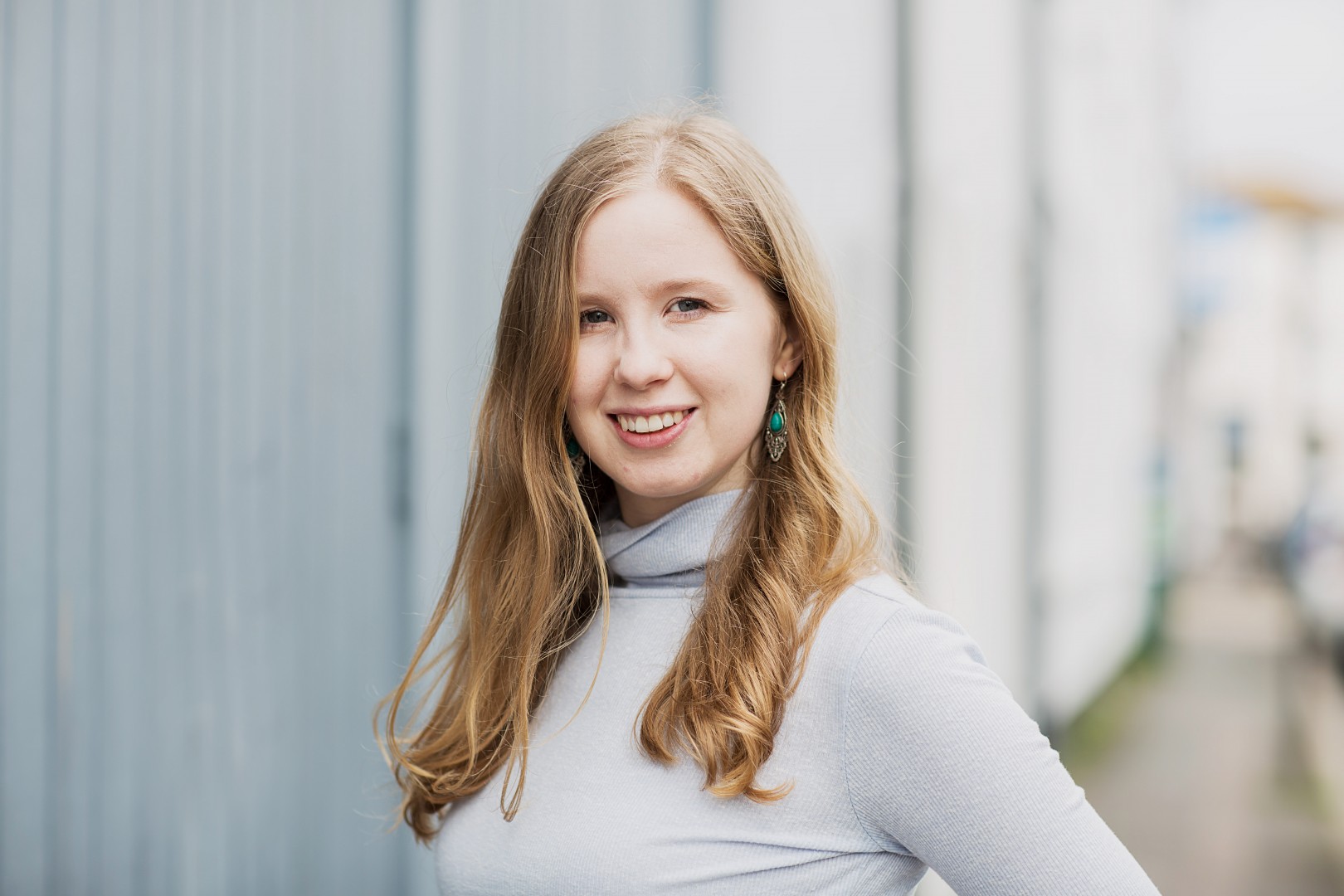 A woman with long blonde hair, wearing a light grey turtleneck and turquoise earrings, smiles outdoors near a lightly colored building.