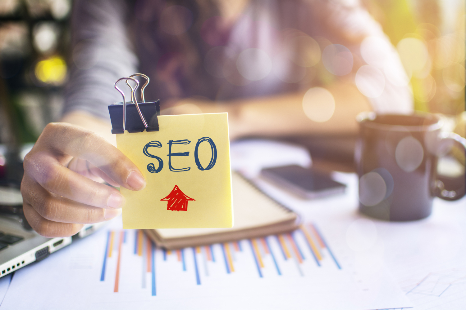 A person holds a yellow sticky note labeled "SEO" with an upward arrow, clipped with a binder clip. In the background, there are charts, a notebook, a smartphone, and a coffee mug on the desk.