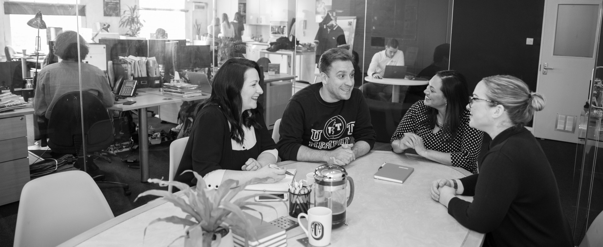Black and white photo of four people sitting around a table in an office, engaged in a conversation, while others work at desks in the background.