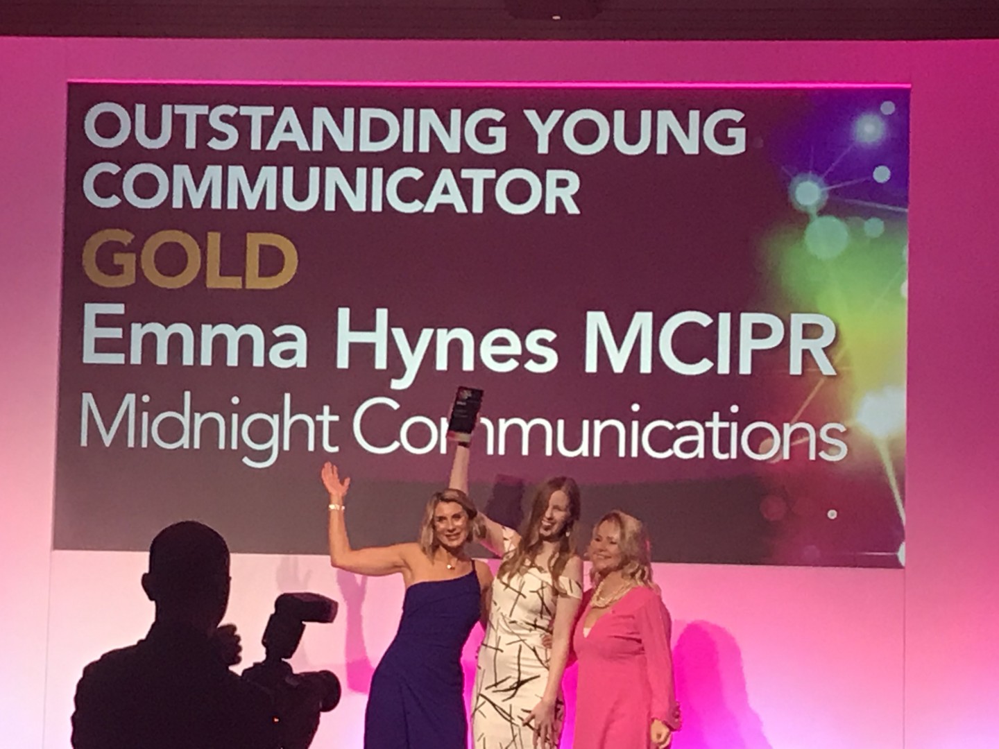 Three women pose on stage in front of an "Outstanding Young Communicator Gold" award sign for Emma Hynes MCIPR of Midnight Communications. One woman holds a trophy, with the other two smiling beside her.