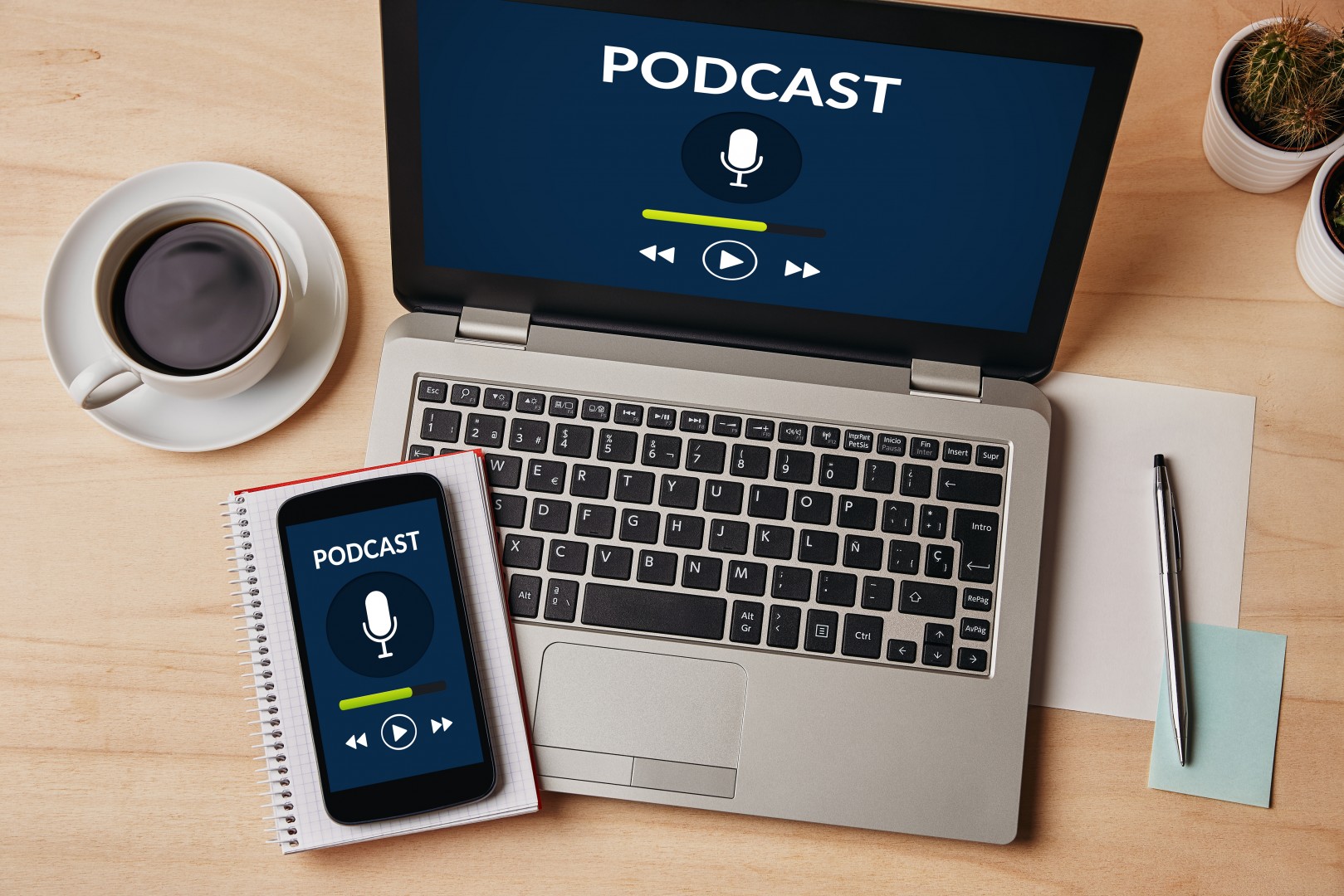 A laptop and smartphone displaying a podcast player screen are placed on a desk next to a cup of coffee, a plant, and a notepad with a pen.