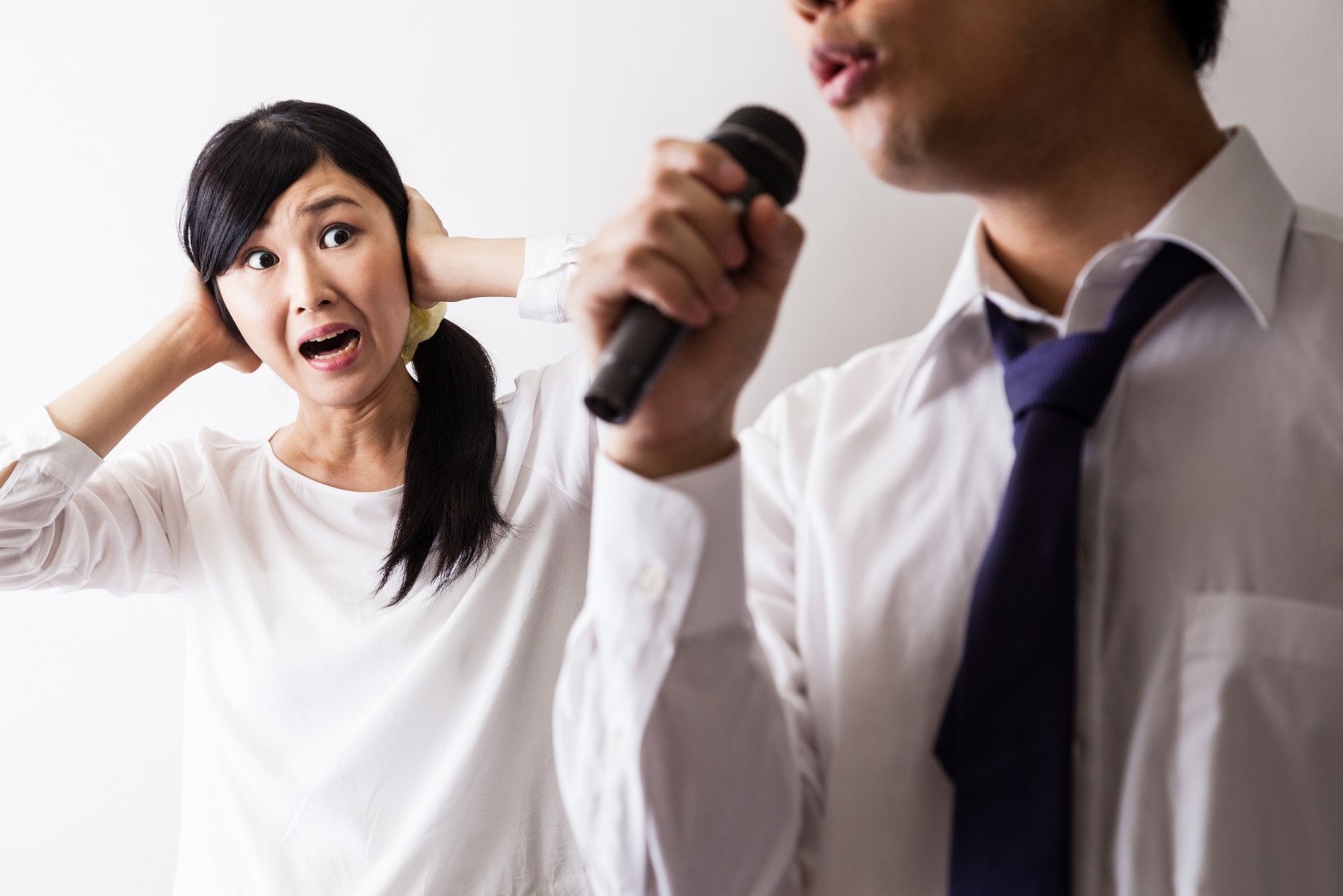 A woman covers her ears with her hands while a man sings into a microphone nearby. Both are wearing white shirts.
