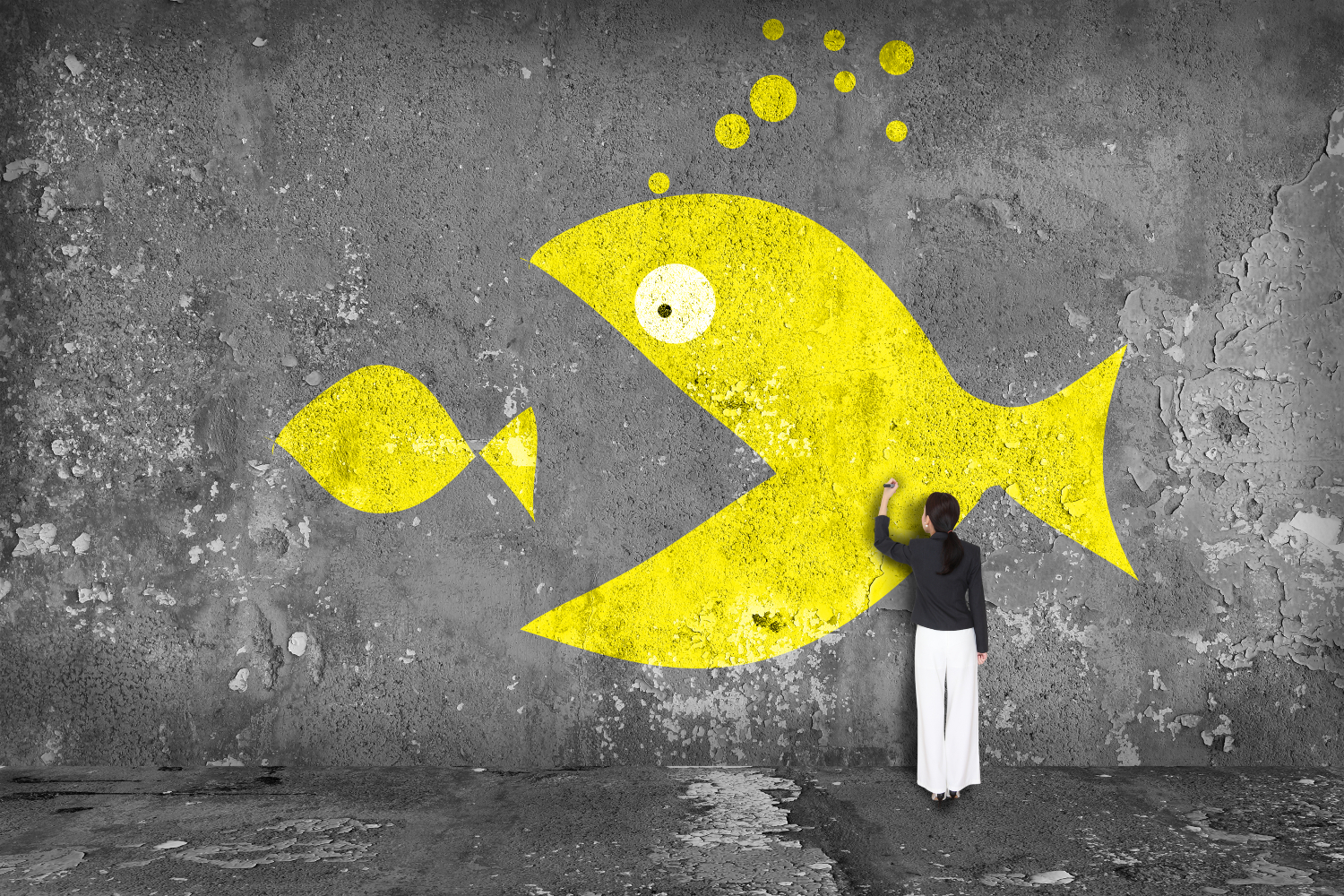 A person in white pants draws a large yellow fish about to eat a smaller yellow fish on a concrete wall.
