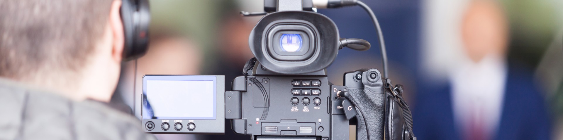 Close-up of a video camera filming an out-of-focus individual in the background. The camera operator is visible on the left side.