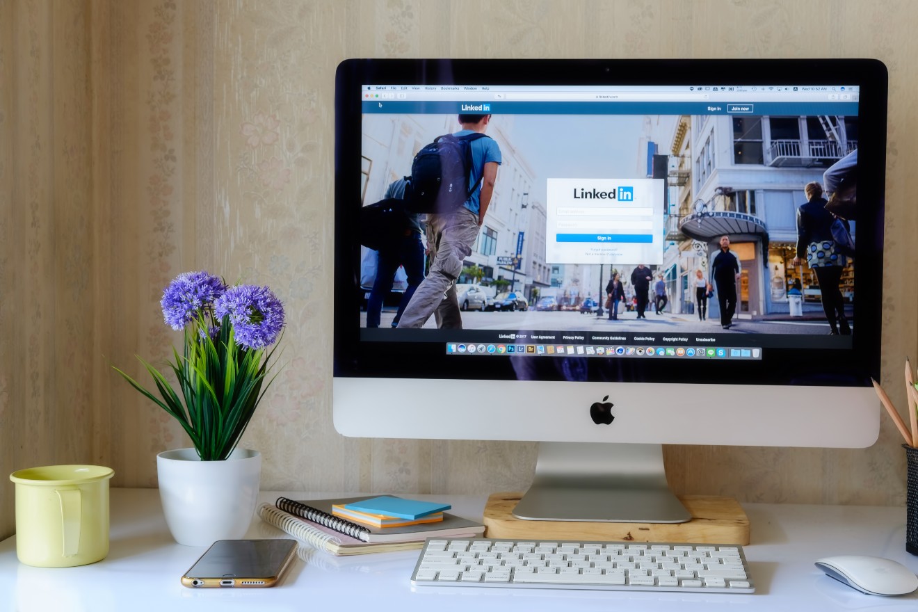 A desktop computer displaying the LinkedIn login page is positioned on a neatly organized desk with notebooks, a smartphone, a plant, a yellow mug, and a pencil holder.