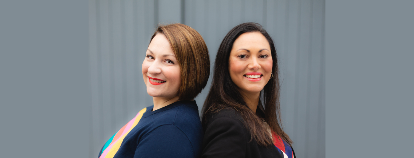 Two women standing back-to-back, smiling, with colorful sashes over their shoulders. The background is a plain, grey vertical panel.