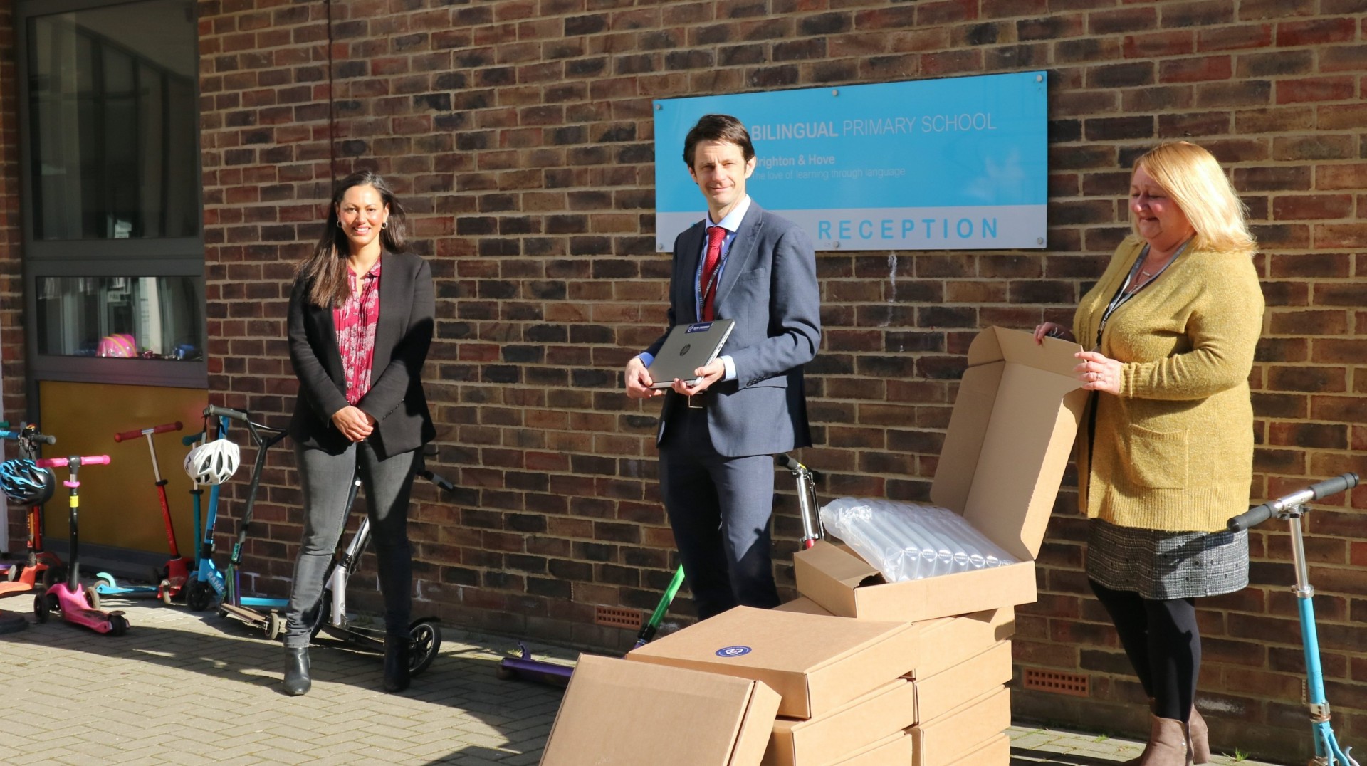Three individuals stand in front of a building with a sign reading "Reception." They are surrounded by boxes and scooters. One person is holding a piece of electronic equipment.