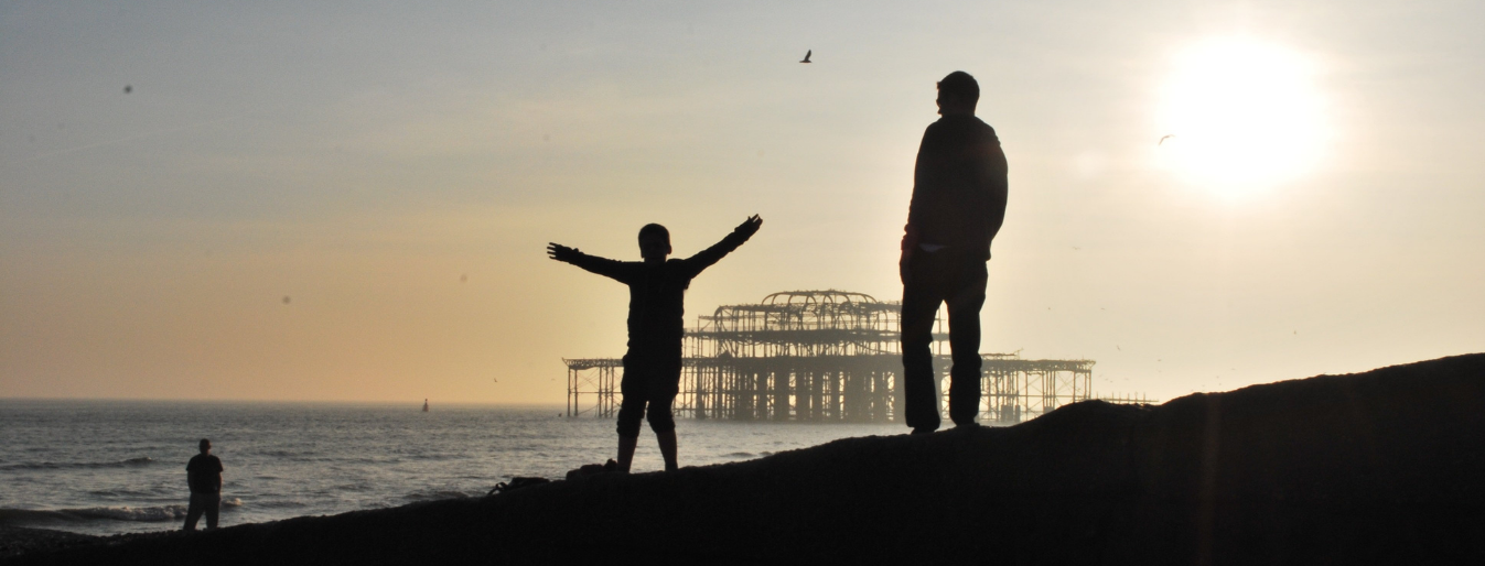 Silhouettes of two individuals standing on the coastline near a ruined pier at sunset. One person has arms outstretched, while another stands with hands in pockets.