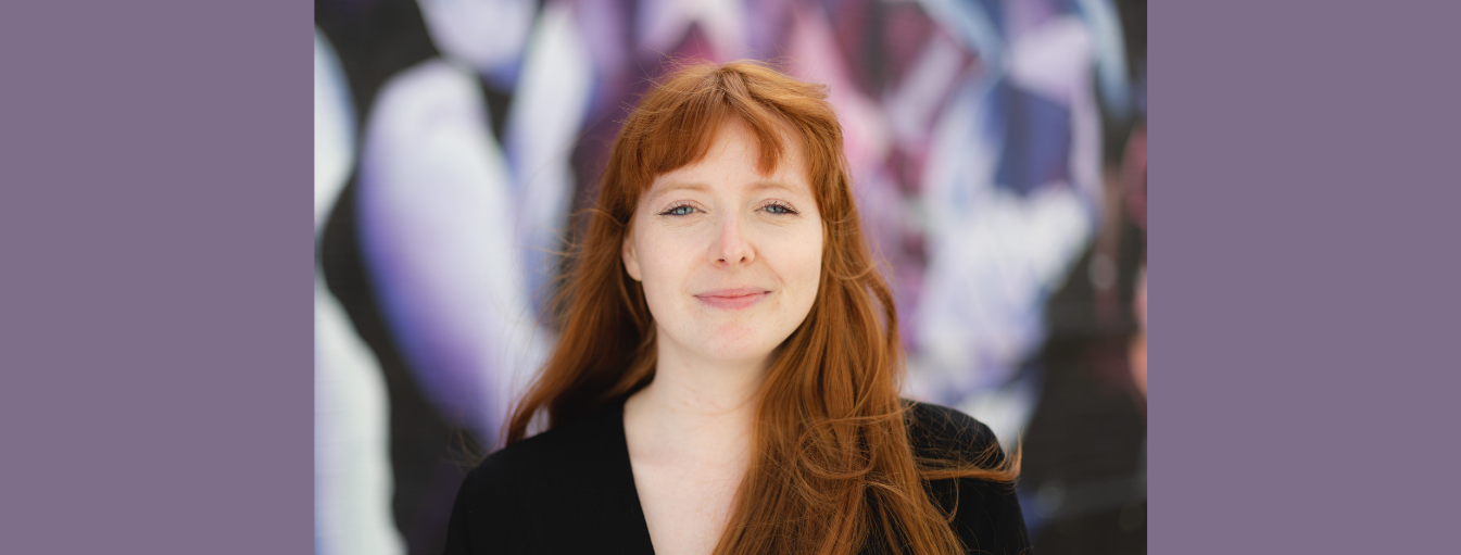 A woman with long red hair and a black top stands in front of a blurred, colorful background.