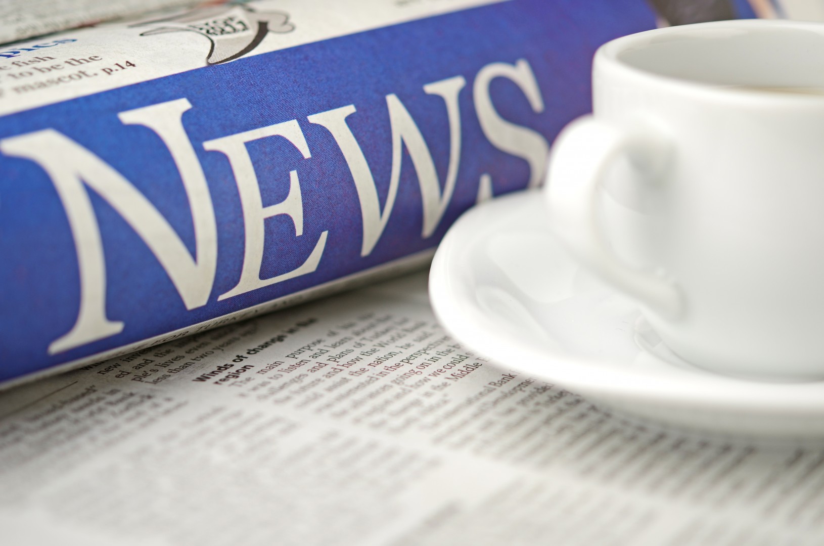 A close-up view of a rolled-up newspaper with the headline "NEWS" in large letters beside a white coffee cup on a saucer, both placed on an open newspaper.