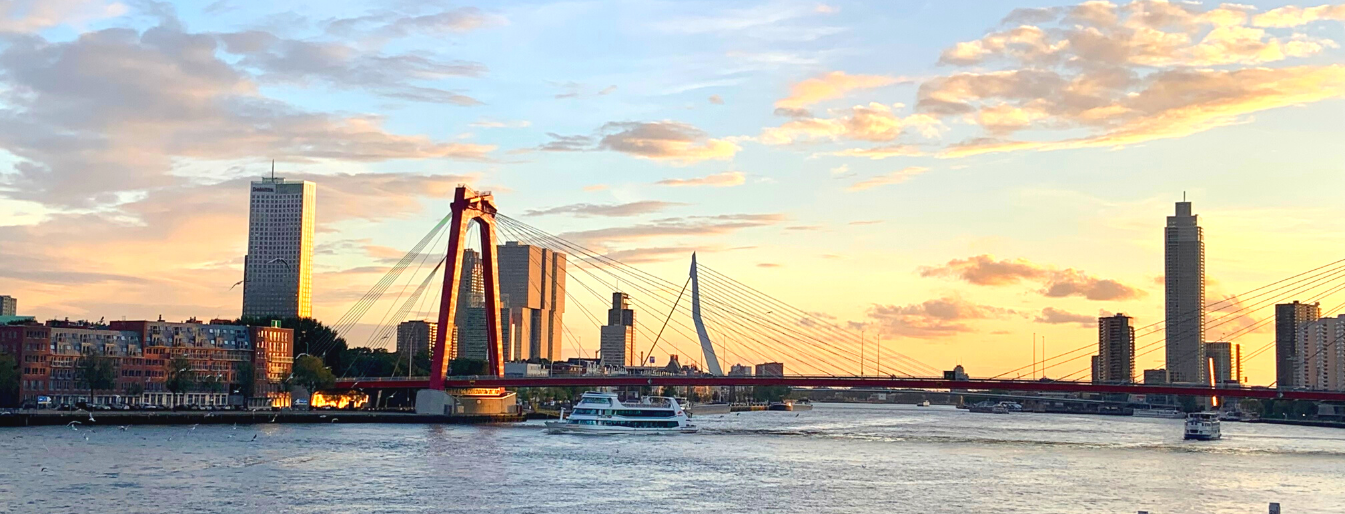 Panoramic view of a river with modern buildings and two bridges at sunset, one with red and the other with white suspension cables, and a boat sailing on the water.