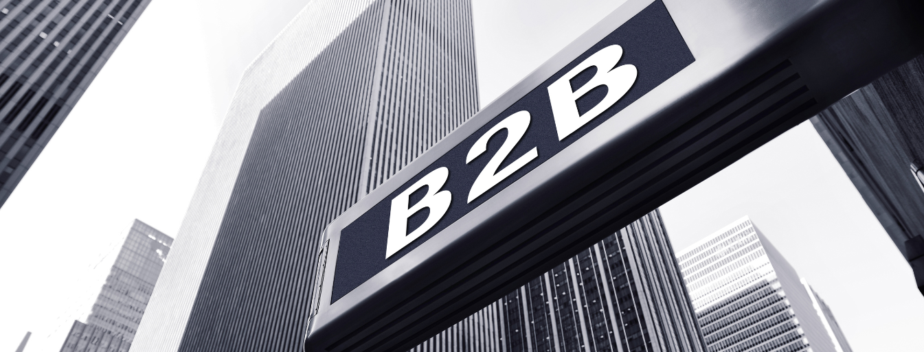 Close-up of a street sign with "B2B" written on it, set against a backdrop of tall modern office buildings in a city.