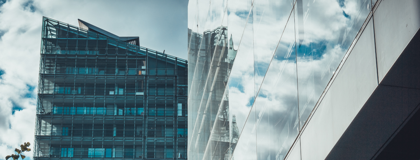 Modern glass building with reflective windows on the left, and another structure with mirrored glass on the right, against a backdrop of a cloudy sky.