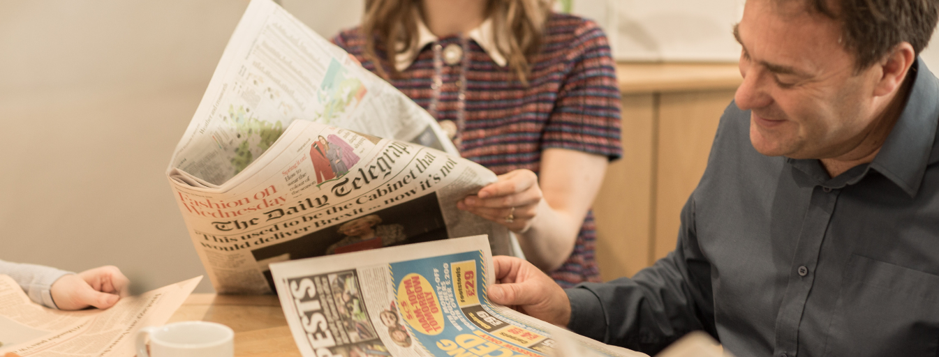 Three people sit at a table reading newspapers. One person holds The Daily Telegraph, another has a different newspaper, and the third person's newspaper is not fully visible. A coffee cup sits on the table.
