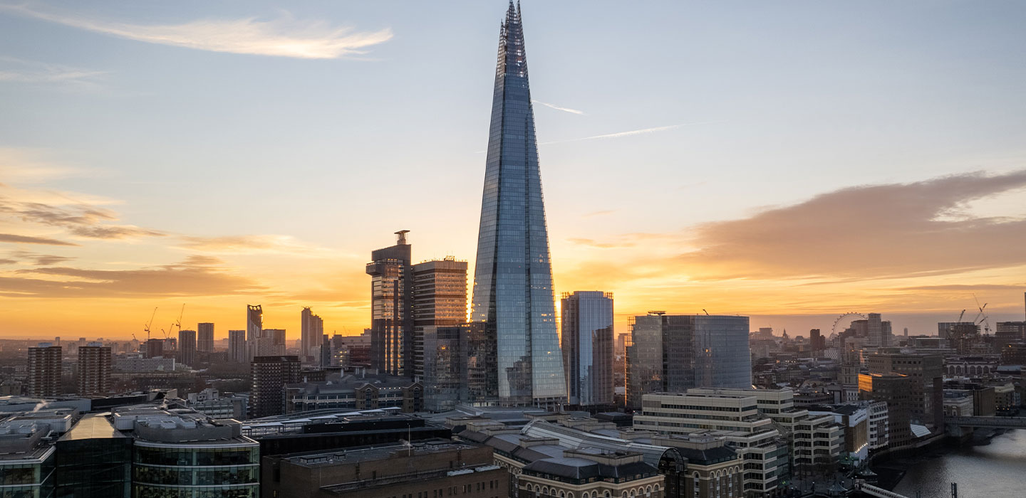 A towering skyscraper with a pointed top, known as The Shard, stands amidst other buildings at sunset in London. The sky is partially cloudy with a warm orange hue.