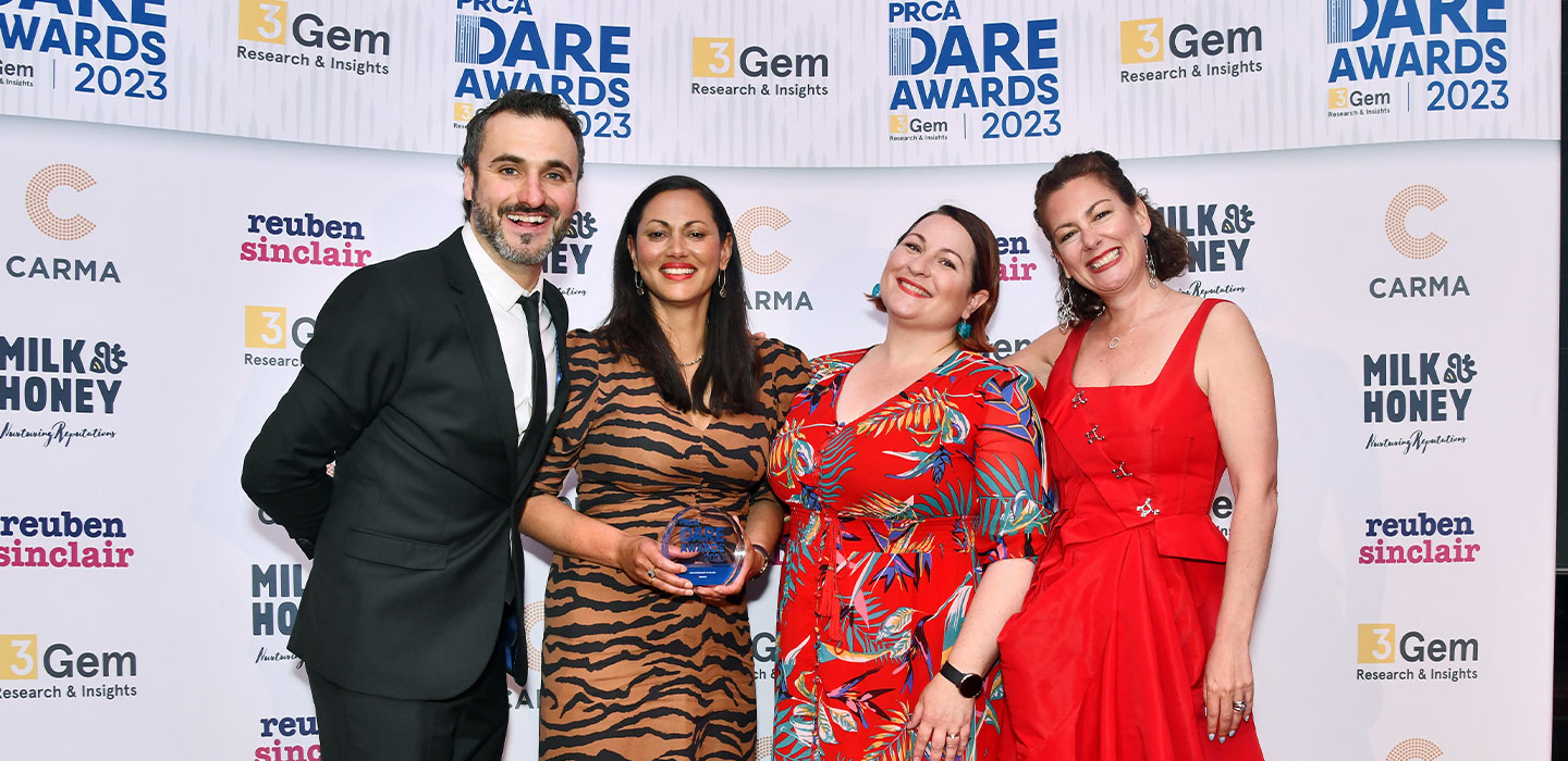 A group of four people stand smiling in front of a PRCA DARE Awards 2023 backdrop, with one person holding an award.
