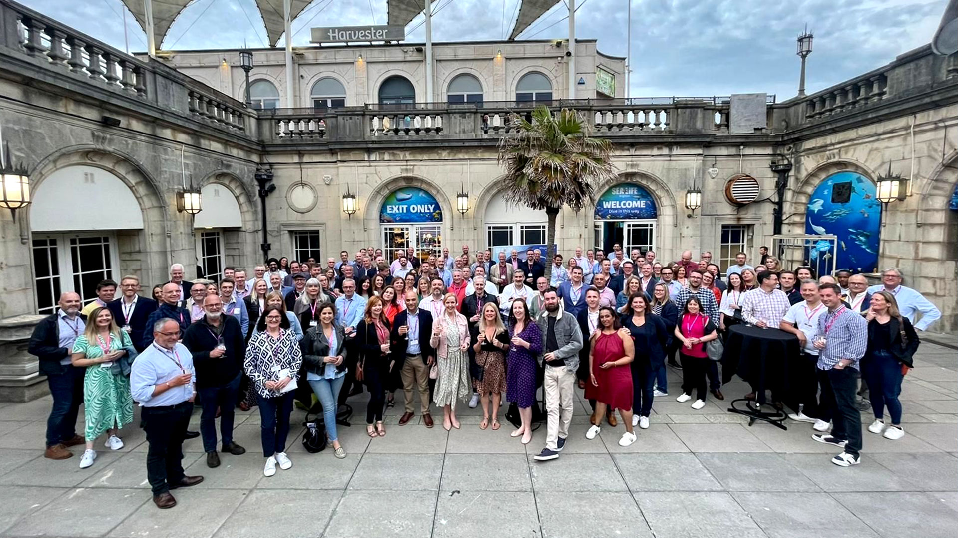 A large group of people stands in an outdoor area lined with stone buildings, gathering for a group photo. The background features a building with "Harvester" signage.