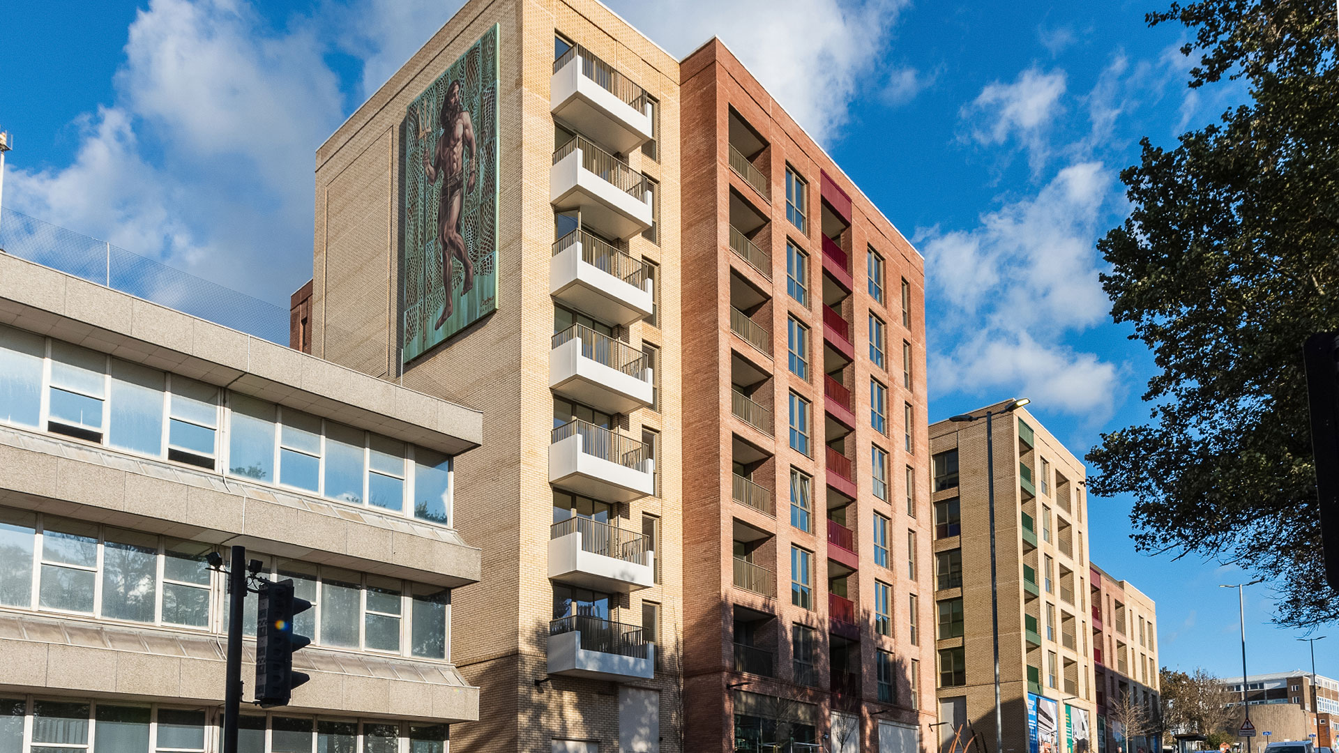 A multi-story building with a large mural on one side and a row of windows on the other is set against a blue sky with scattered clouds.