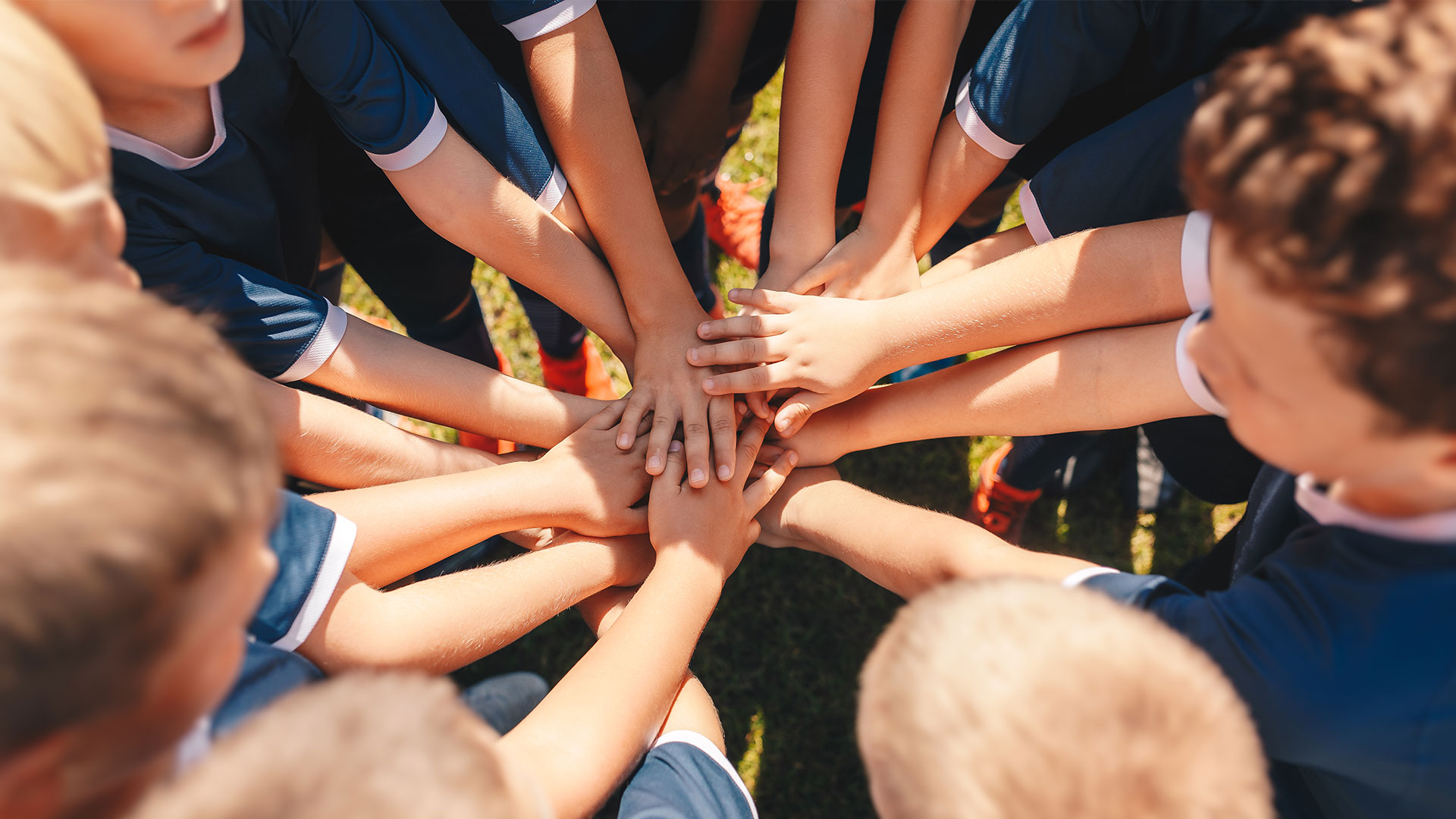 A group of young people in sports uniforms stand in a huddle, placing their hands together in the center.