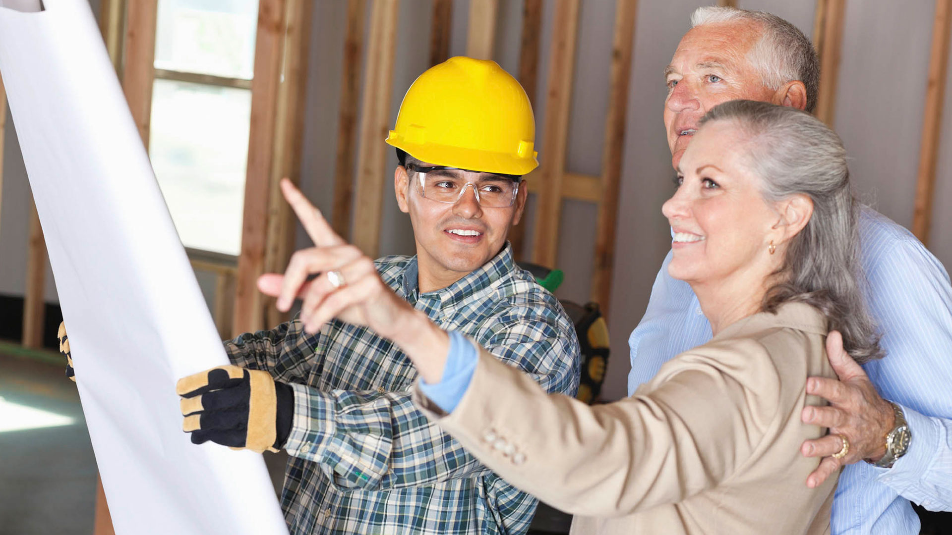 A construction worker in a yellow hard hat shows building plans to an elderly couple inside a partially constructed house.