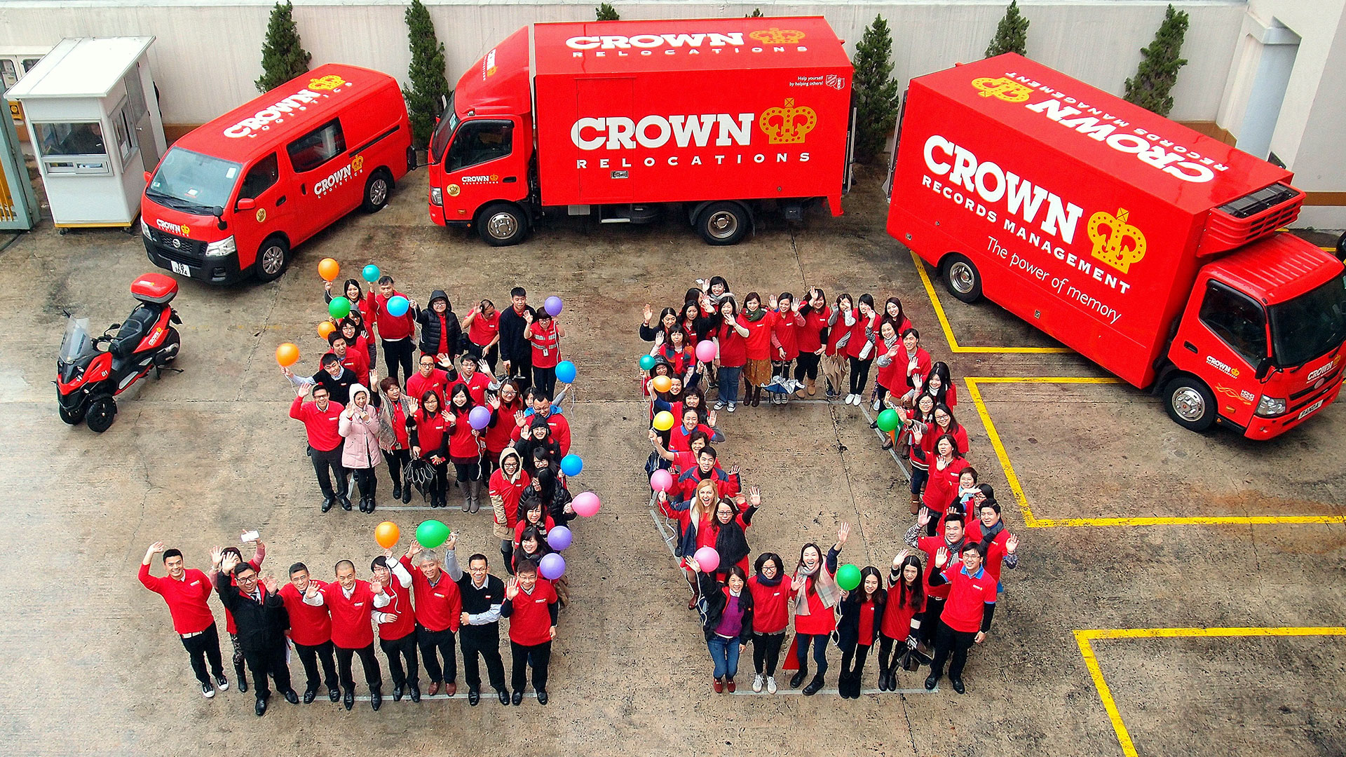 A group of people in red shirts stand arranged to form the number '60' surrounded by red Crown Relocations trucks and vans. Some individuals hold colorful balloons.