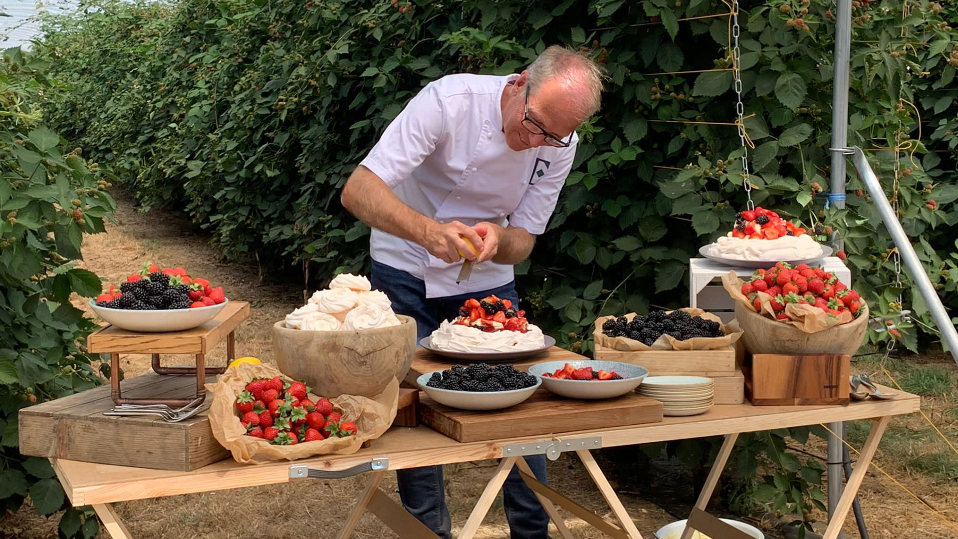 A man in a white chef's jacket is decorating a dessert with fruit at an outdoor table filled with various berries and meringues.