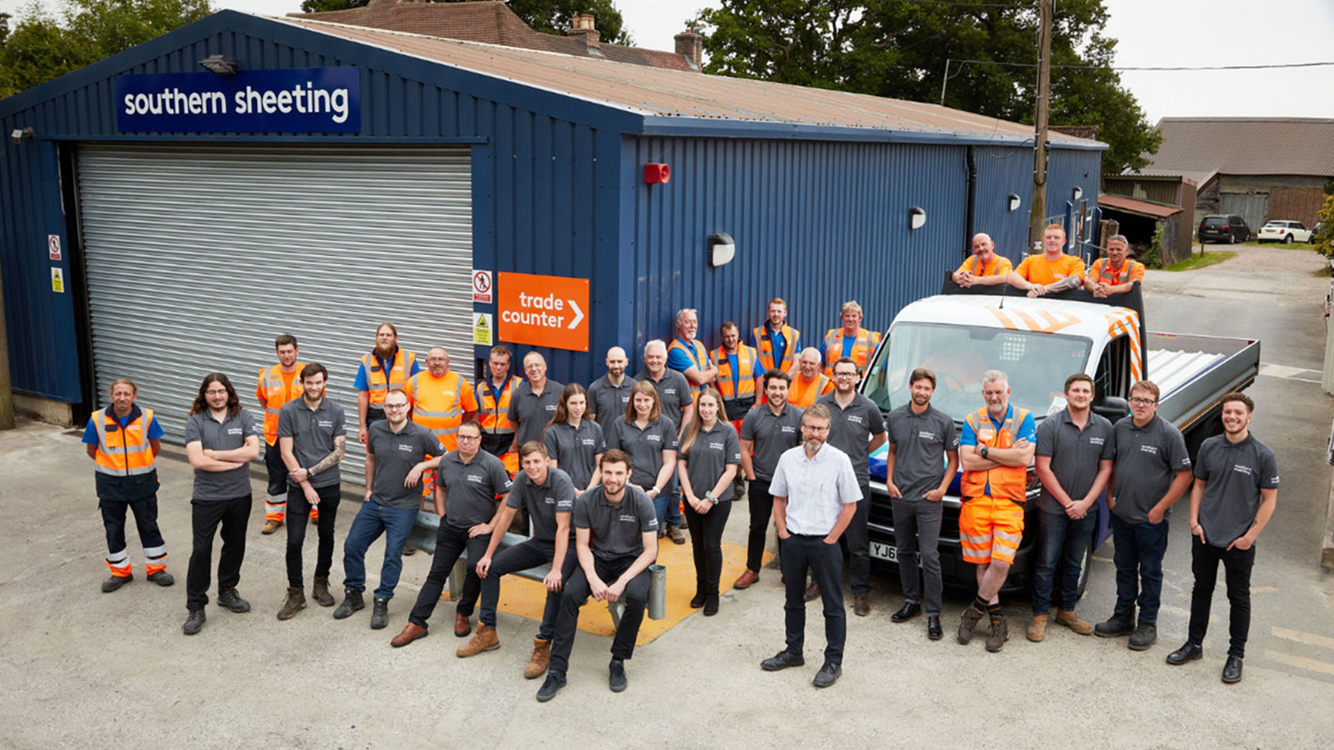 A group of employees wearing safety vests and company shirts gather in front of a blue industrial building labeled "southern sheeting" with a sign indicating a trade counter. A truck is parked nearby.