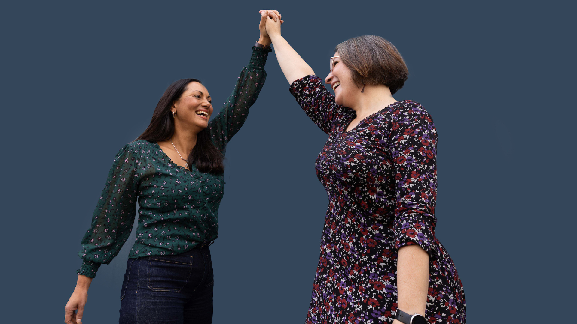 Two women smiling and holding hands up, wearing patterned tops, against a solid dark blue background.