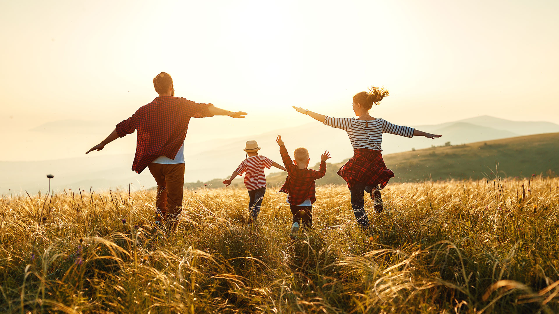 Four people in casual clothes run joyfully through a grassy field at sunset, with arms outstretched. Hills are visible in the background under a clear sky.