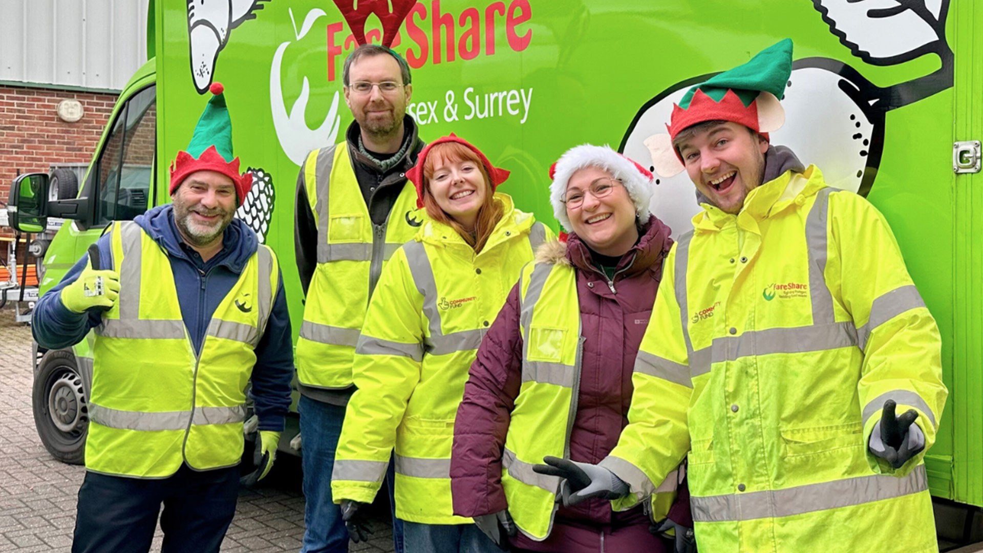 Five people wearing festive hats and high-visibility jackets smile and gesture in front of a green van with a "FareShare Sussex & Surrey" logo.