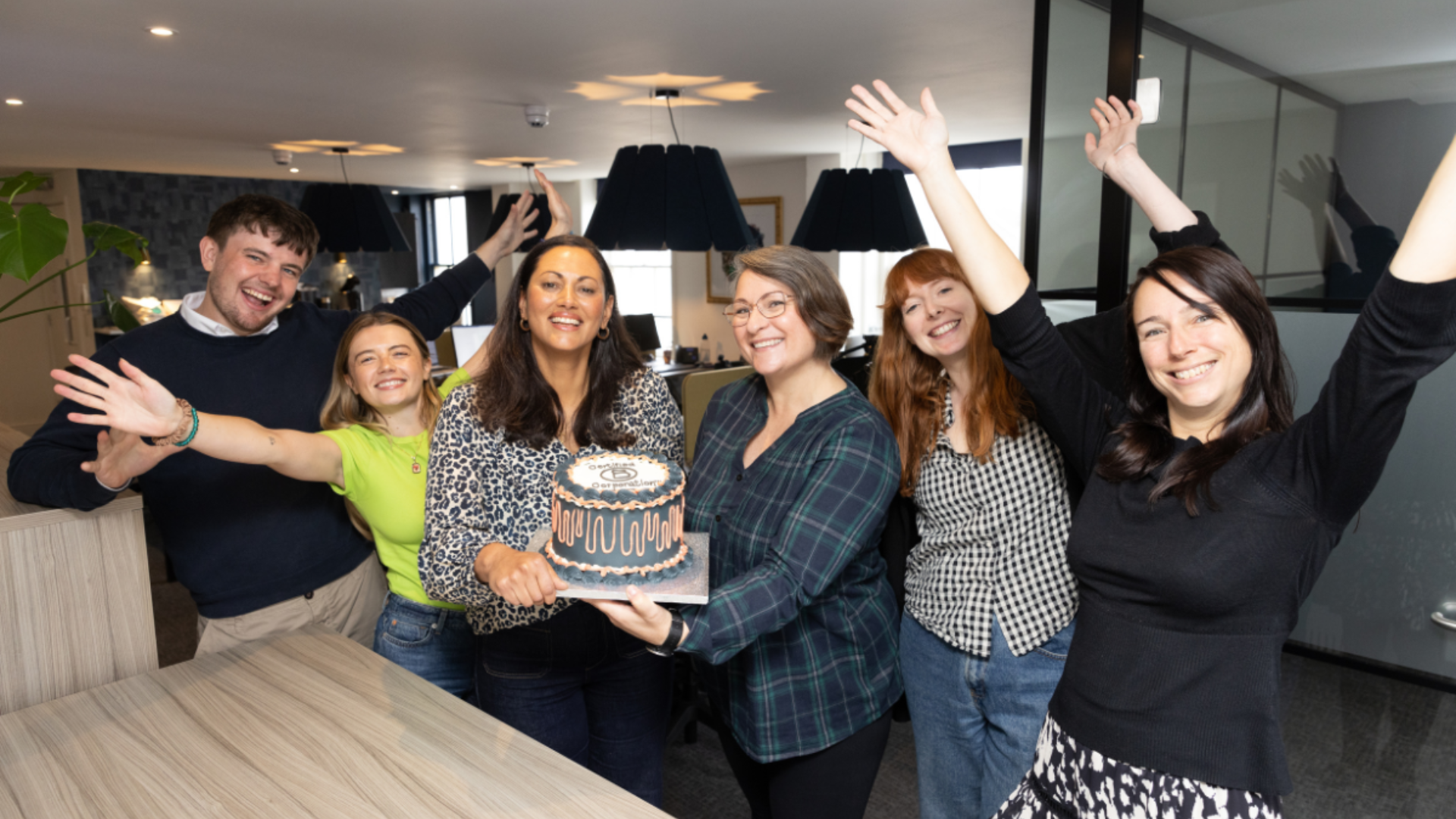 Six people stand indoors, smiling and raising their arms, while two of them hold a cake decorated with "25" on top, celebrating an anniversary or milestone.